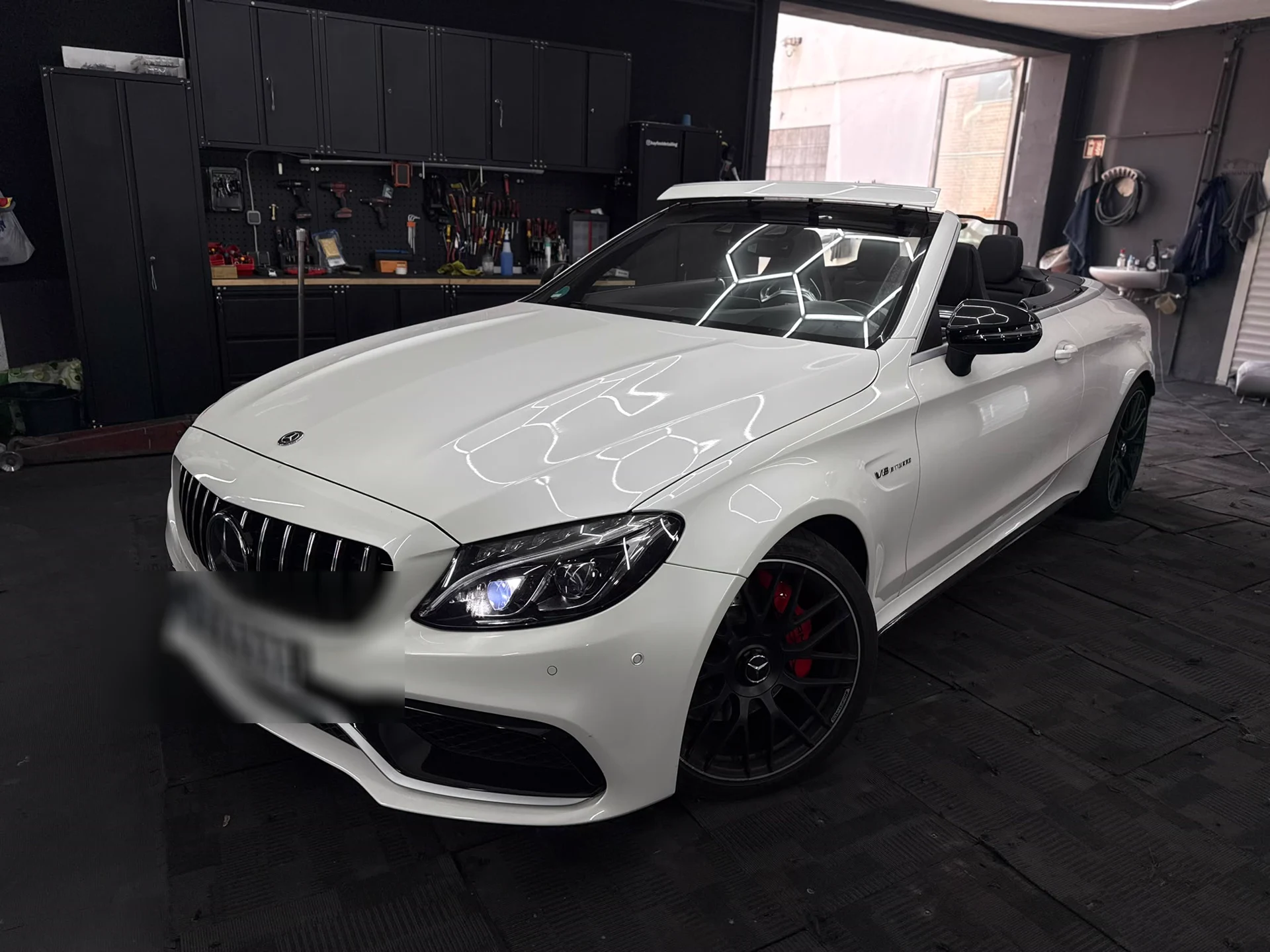 Weißes Mercedes-AMG Cabrio in der Halle, die Hexagon-Deckenbeleuchtung spiegelt sich auf Haube und Kotflügel